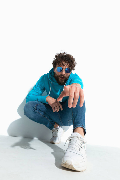 attractive casual guy with cool style is pointing at the camera while squatting on white background - Photo, Image