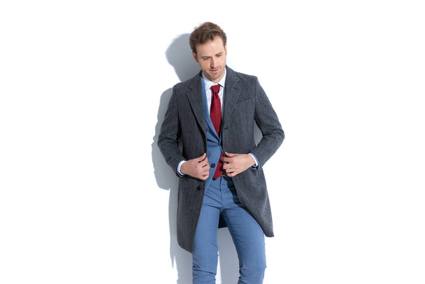 attraktiver Geschäftsmann, der seinen Mantel arrangiert, nach unten schaut und vor weißem Hintergrund posiert - Foto, Bild
