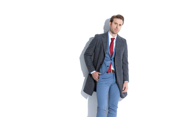 cool fashion model wearing a nice blue suit and a coat over and holding one hand in pocket  - Fotografie, Obrázek