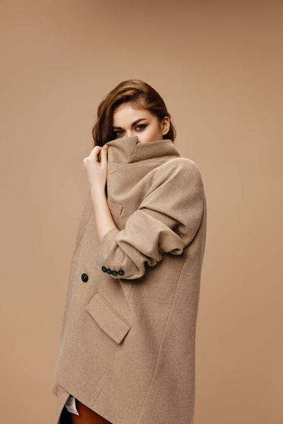 woman in coat covers her face with collar and beige background model - Foto, afbeelding