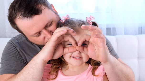 Pieni tytär 3-vuotias ja isä pitävät hauskaa kotona, tekevät hauskoja kasvoja, tekevät silmälaseja sormillaan, kuten lasit katselevat kameraa kiikarien läpi. Pidä hauskaa lapsen kanssa, ilmaiseksi - Valokuva, kuva