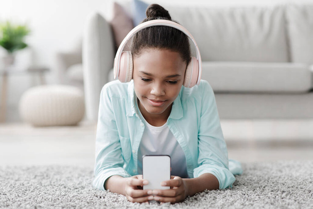 Сконцентрированная улыбающаяся американка-подросток учится, смотрит онлайн урок по телефону, лежит на полу - Фото, изображение