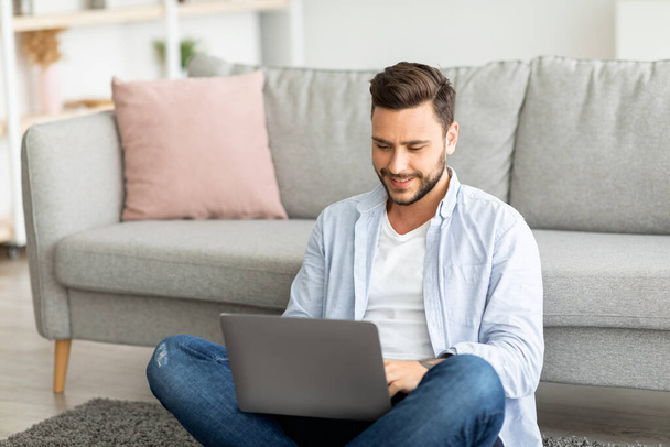Mosolygó Y-generációs férfi dolgozik otthonról, ül a padlón, és használja laptop, szabad hely - Fotó, kép