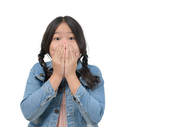 Překvapená holčička v džínové bundě zakrývající ústa a při pohledu na kameru izolované na bílém pozadí, šokovaný emoční koncept - Fotografie, Obrázek
