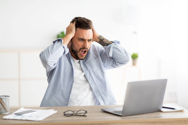 Homme en colère ayant le logiciel, erreur de site Web, regardant l'écran d'ordinateur portable et gestuelle, assis au lieu de travail à l'intérieur de la maison - Photo, image