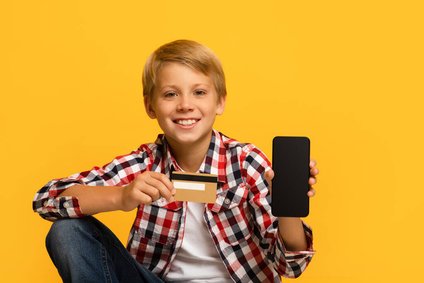 Vidám jóképű tini szőke fiú tanuló show okostelefon üres képernyő és hitelkártya - Fotó, kép