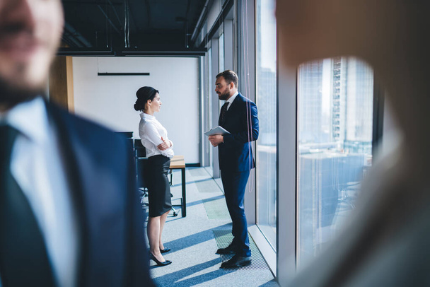 Side view of female executive having conversation with male employee holding tablet while standing near window of modern office building with blurred people in foreground - 写真・画像