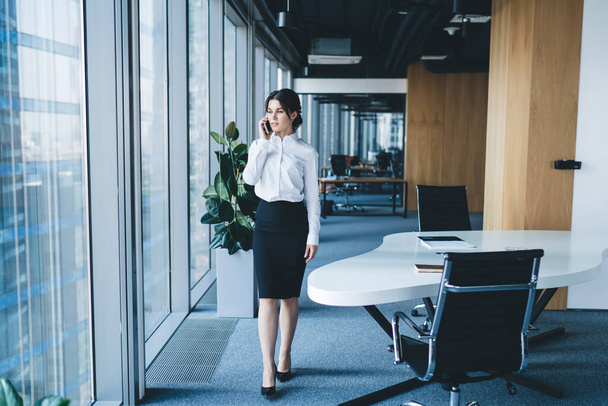 Serious elegant woman in stylish clothes standing by table in open space office while talking on smartphone near panoramic window - Фото, зображення