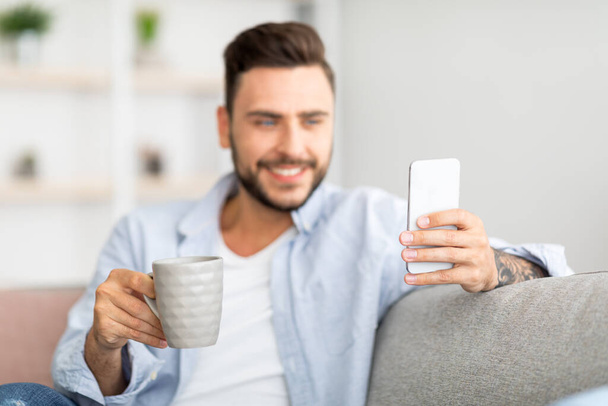 Online life concept. Excited millennial man holding modern smartphone and cup of coffee at home, free space - Фото, зображення