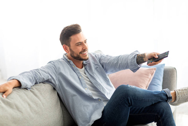 Quédate en casa entretenimientos. Hombre relajado con mando a distancia viendo la televisión en el sofá en la sala de estar, espacio libre - Foto, imagen