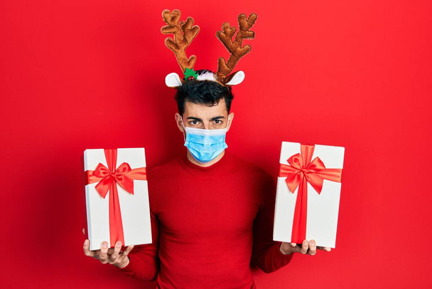 Jeune homme hispanique portant de jolies cornes de rennes de Noël et masque médical tenant des cadeaux déprimés et inquiets pour la détresse, pleurant en colère et effrayés. triste expression.  - Photo, image