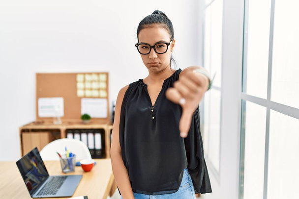 オフィスで働く若いヒスパニック系のビジネスマンの女性は、親指ダウンジェスチャーで拒絶と否定を示す不満と怒りを見ています。悪い表現.  - 写真・画像