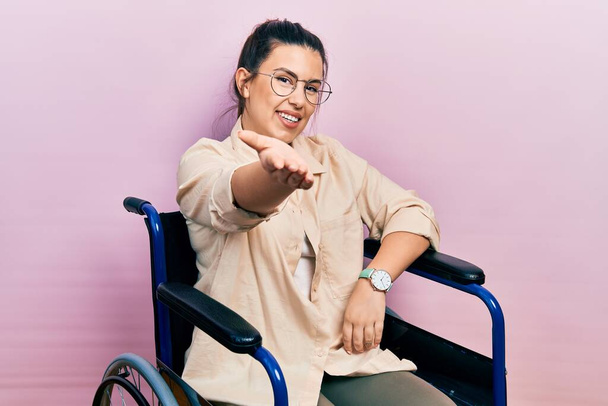Young hispanic woman sitting on wheelchair smiling cheerful offering palm hand giving assistance and acceptance.  - Photo, Image