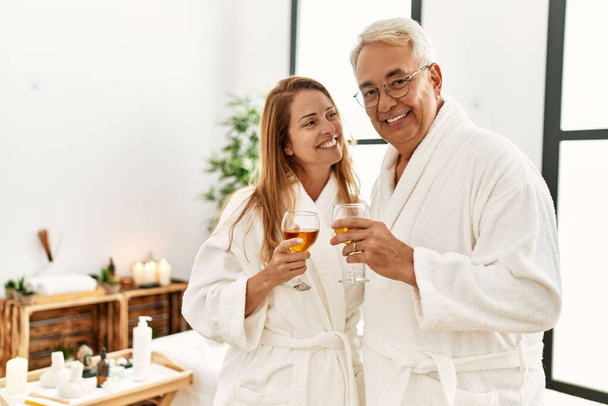 Latynoska para w średnim wieku uśmiechnięta szczęśliwa pijąc szampana w centrum odnowy biologicznej. - Zdjęcie, obraz