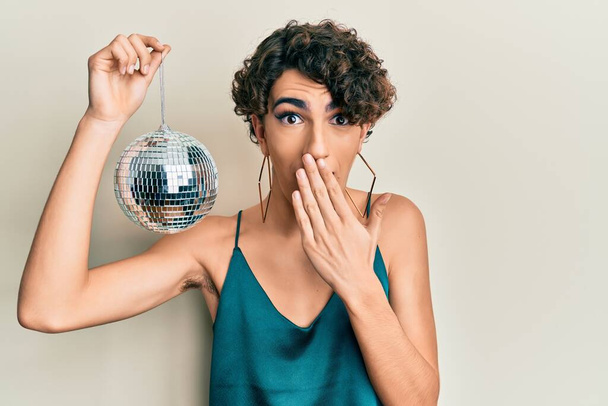 Young hispanic gay holding shiny disco ball covering mouth with hand, shocked and afraid for mistake. surprised expression  - Foto, Imagem