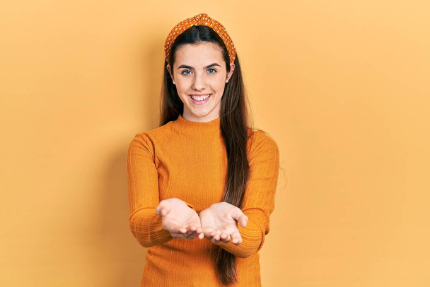 Młoda brunetka nastolatka ubrana w luźny żółty sweter uśmiechnięta dłońmi razem otrzymująca lub dająca gest. trzymać i chronić  - Zdjęcie, obraz