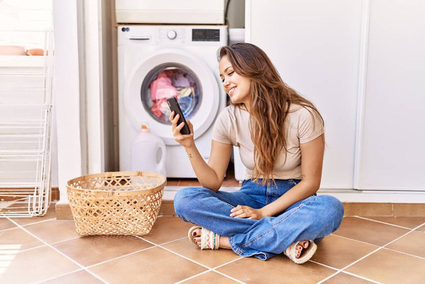 Joven chica hispana usando smartphone esperando la lavadora en casa - Foto, Imagen