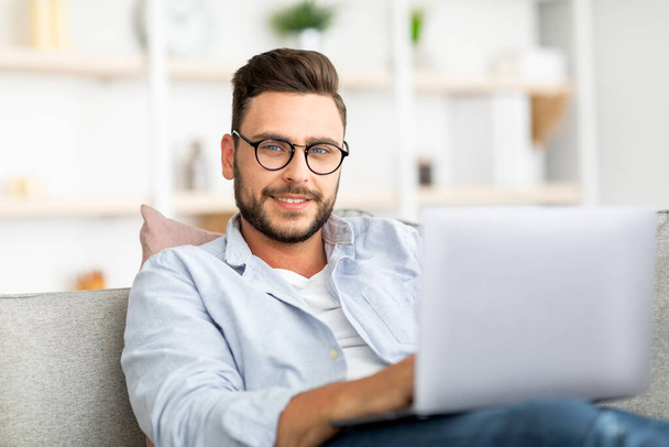 Gözlüklü yakışıklı adam evde dizüstü bilgisayar kullanıyor, koltukta oturuyor, evden çalışıyor, e-posta yazıyor ya da programlıyor. - Fotoğraf, Görsel