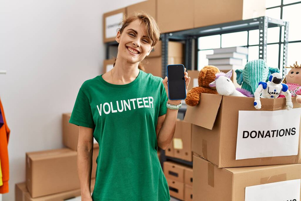 Nuori valkoihoinen vapaaehtoinen nainen hymyilee onnellinen osoittaa vapaaehtoistyötä sovellus hyväntekeväisyyskeskuksessa. - Valokuva, kuva