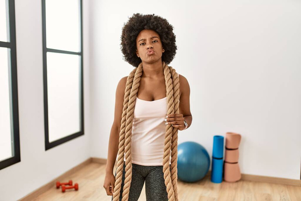 Νεαρή Αφροαμερικανή γυναίκα με αφρο μαλλιά στο γυμναστήριο προπόνηση με σχοινιά μάχης κοιτάζοντας την κάμερα φυσάει ένα φιλί στον αέρα είναι όμορφη και σέξι. έκφραση αγάπης.  - Φωτογραφία, εικόνα