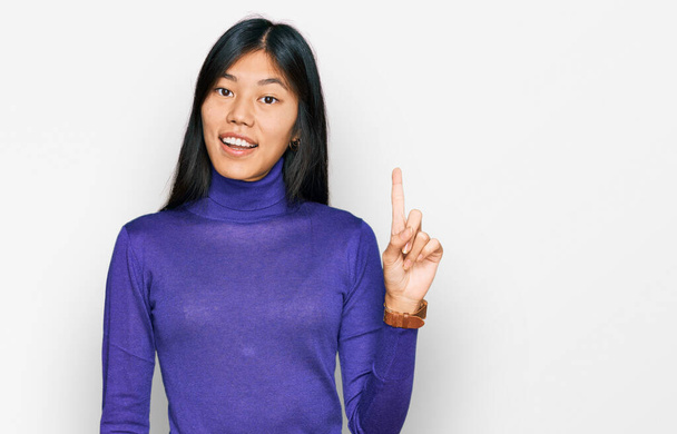 Mooie jonge Aziatische vrouw draagt casual kleding wijzen vinger omhoog met een succesvol idee. opgewonden en gelukkig. Nummer een.  - Foto, afbeelding