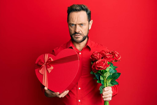Middle age man holding anniversary present and bouquet of flowers clueless and confused expression. doubt concept.  - Photo, Image