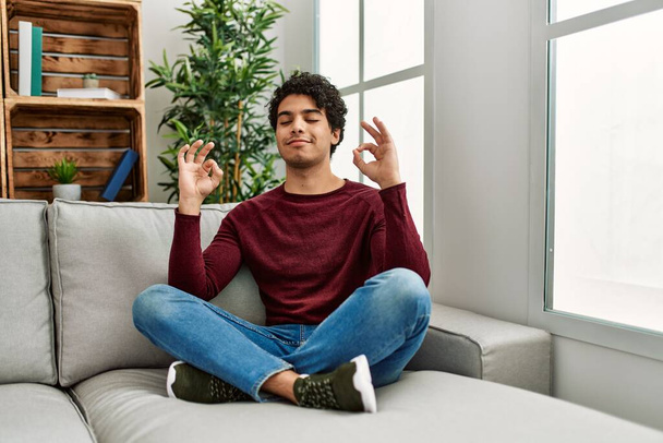 Fiatal spanyol férfi meditáció jóga ül a kanapén otthon. - Fotó, kép