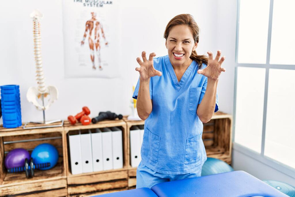 Hispanische Physiotherapeutin mittleren Alters, die in der Schmerzklinik arbeitet, lächelt lustig und macht Krallen-Geste als Katze, aggressiver und sexy Ausdruck  - Foto, Bild