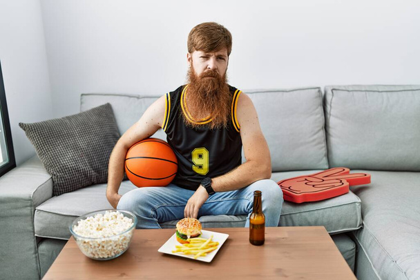 Kaukasischer Mann mit langem Bart, Basketballball in der Hand, der das Fernsehspiel anfeuert, sieht schläfrig und müde aus, erschöpft vor Müdigkeit und Kater, faule Augen am Morgen.  - Foto, Bild