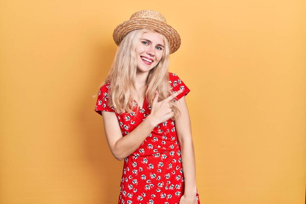 piękna kaukaska kobieta z blond włosy noszenie letni kapelusz wesoły z uśmiechem twarzy wskazując z ręki i palca do góry na bok z szczęśliwy i naturalny wyraz twarzy  - Zdjęcie, obraz