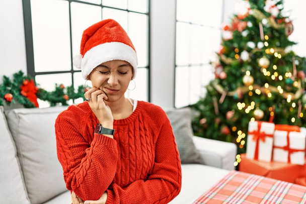Mujer hispana joven con el pelo corto con sombrero de Navidad sentado en el sofá tocando la boca con la mano con expresión dolorosa debido a dolor de muelas o enfermedad dental en los dientes. dentista  - Foto, Imagen