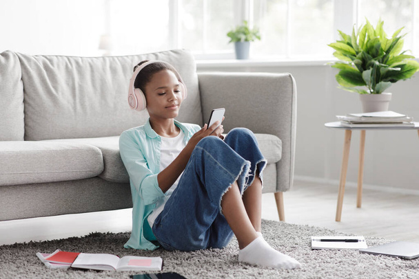 Šťastná mladá černoška vychutnat oblíbenou hudbu, sledovat video na telefonu, sedí na podlaze, doma interiér - Fotografie, Obrázek