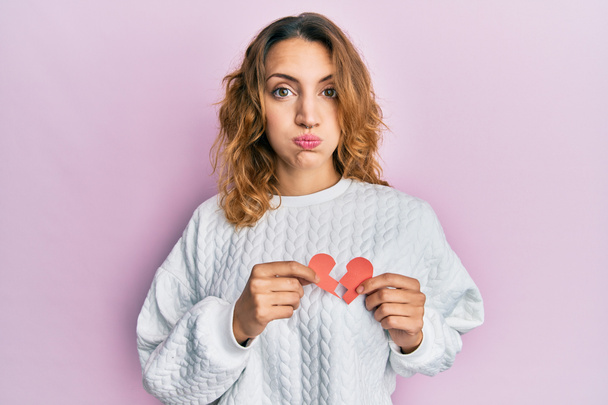 Young caucasian woman holding broken heart paper shape puffing cheeks with funny face. mouth inflated with air, catching air.  - Foto, immagini