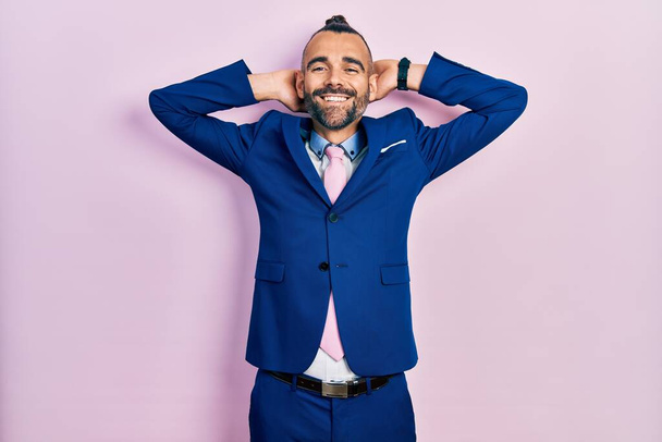 Νεαρός Ισπανός που φοράει κοστούμι και γραβάτα χαλαρώνοντας και τεντώνοντας, χέρια και χέρια πίσω από το κεφάλι και το λαιμό χαμογελώντας χαρούμενος  - Φωτογραφία, εικόνα