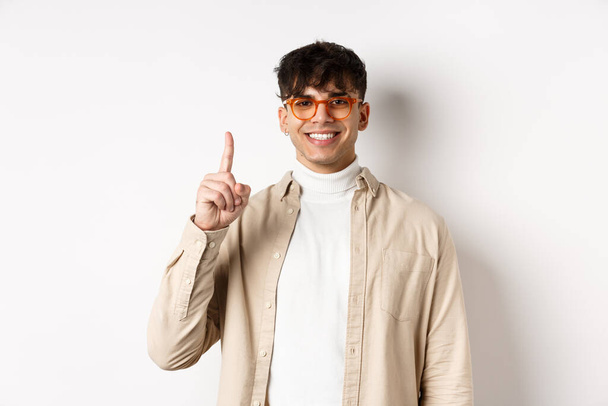 Guapo caucásico chico en gafas mostrando un dedo y sonriendo, de pie sobre fondo blanco - Foto, imagen