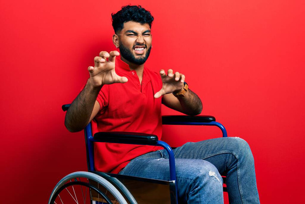 Hombre árabe con barba sentado en silla de ruedas sonriendo divertido haciendo gesto de garra como gato, expresión agresiva y sexy  - Foto, Imagen