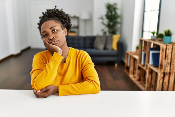 Jeune femme afro-américaine portant des vêtements décontractés assis sur la table à la maison pensant fatigué et ennuyé avec des problèmes de dépression avec les bras croisés.  - Photo, image