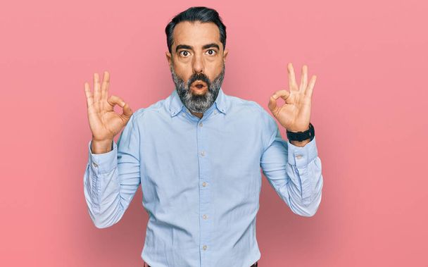 Hombre de mediana edad con barba vistiendo camisa de negocios que parece sorprendido y sorprendido haciendo símbolo de aprobación bien con los dedos. expresión loca  - Foto, imagen