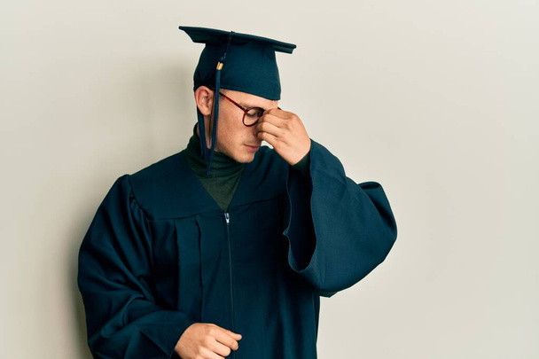 Young caucasian man wearing graduation cap and ceremony robe tired rubbing nose and eyes feeling fatigue and headache. stress and frustration concept.  - Foto, Imagen