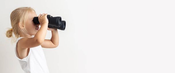 Smiling blonde child looks through binoculars on a light background in the studio. Banner. - Foto, Imagem