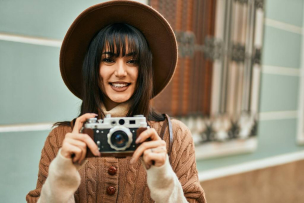 Μελαχρινή γυναίκα φοράει χειμερινό καπέλο χαμογελώντας χρησιμοποιώντας vintage κάμερα σε εξωτερικούς χώρους στην πόλη - Φωτογραφία, εικόνα