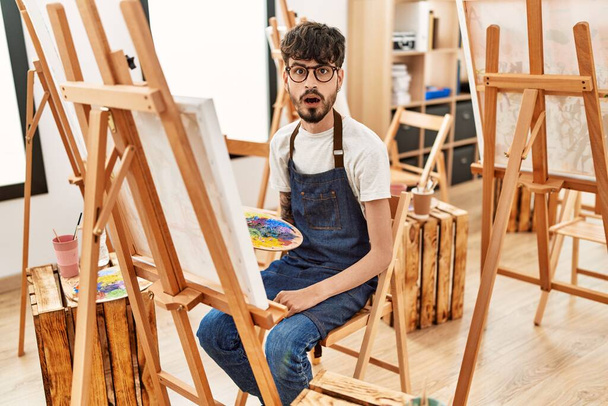 Homme hispanique avec barbe au studio d'art effrayé et étonné avec la bouche ouverte pour la surprise, visage incrédule  - Photo, image