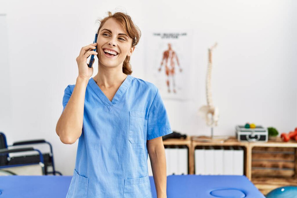 Junge kaukasische Physiotherapeutin lächelt fröhlich auf dem Smartphone in der Klinik. - Foto, Bild