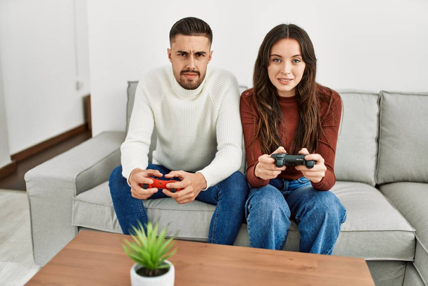 Νεαρό ζευγάρι ισπανόφωνων που παίζει βιντεοπαιχνίδι κάθεται στον καναπέ στο σπίτι. - Φωτογραφία, εικόνα