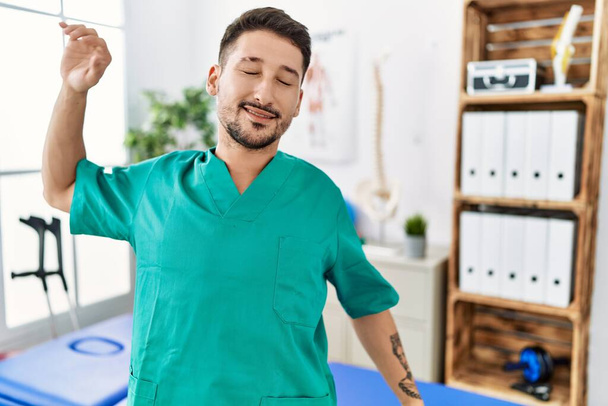 Young physiotherapist man working at pain recovery clinic stretching back, tired and relaxed, sleepy and yawning for early morning  - Foto, Imagem