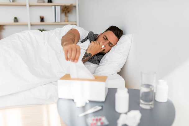 Erkältung, Grippe, Coronavirus. Kranker nimmt Servietten aus Gewebebox und niest laufende Nase, liegt im Bett - Foto, Bild