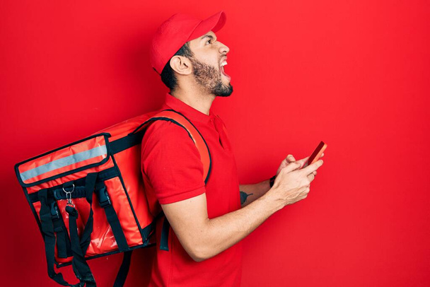 Hispanic man with beard wearing delivery package backpack using smartphone angry and mad screaming frustrated and furious, shouting with anger looking up.  - Photo, image