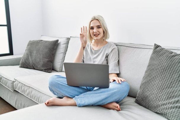 Jeune femme caucasienne utilisant ordinateur portable à la maison assis sur le canapé montrant et pointant vers le haut avec les doigts numéro trois tout en souriant confiant et heureux.  - Photo, image