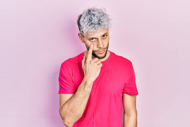Joven hombre hispano con el pelo teñido moderno con camiseta rosa casual apuntando al ojo observándote gesto, expresión sospechosa  - Foto, Imagen