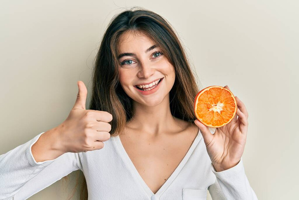 Young caucasian woman holding fresh slice of orange smiling happy and positive, thumb up doing excellent and approval sign  - Photo, image